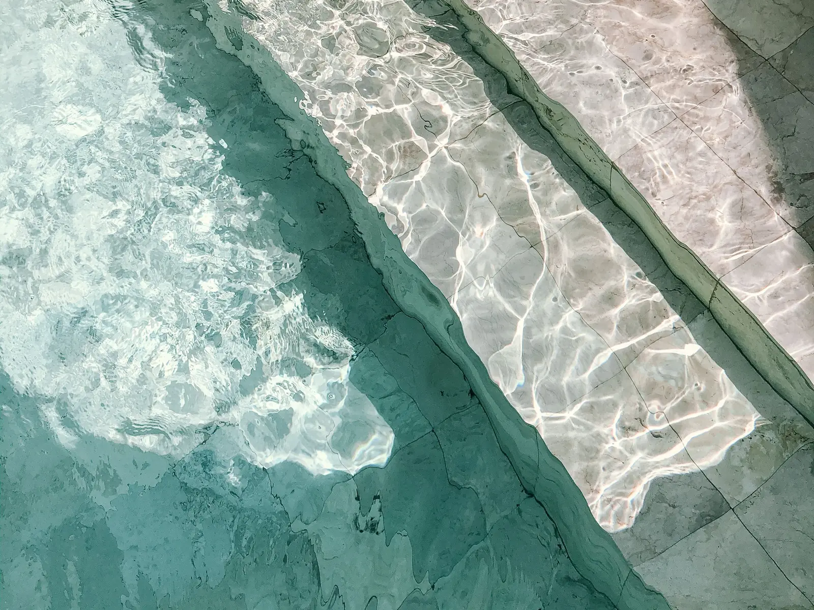 Sunlight rippling across the turquoise water and travertine steps of a pool
