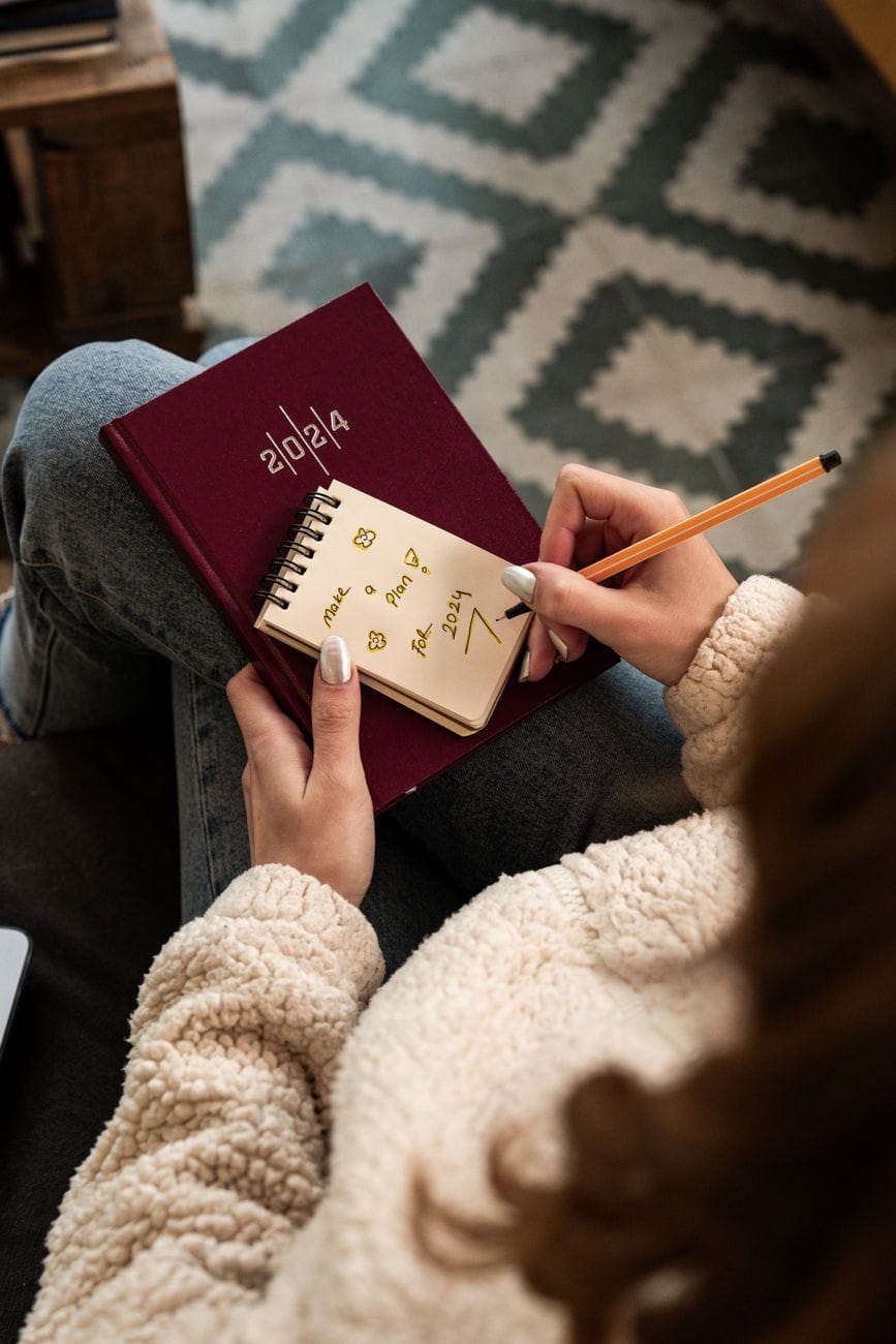 A hand writing 'make a plan' in a small notebook on a cosy rug