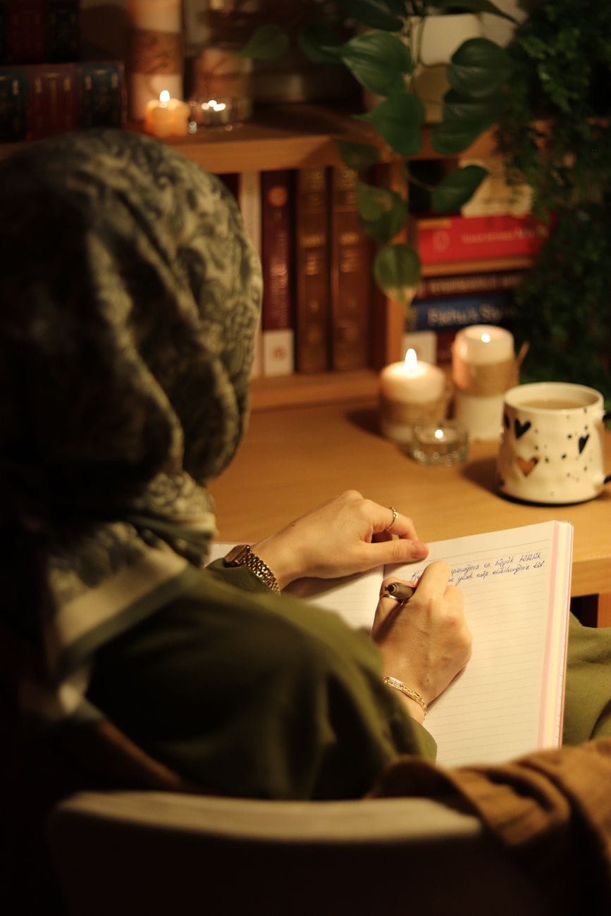 A woman writing in a notebook by candlelight surrounded by books and plants