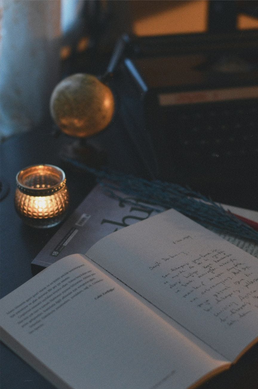 An open handwritten journal beside a lit candle and a small brass globe
